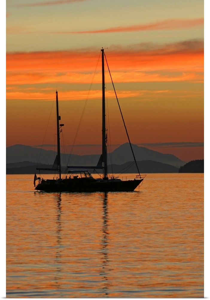 Beautiful photograph of a sail boat sitting in the water with mountains in the background as the sun sets and lights up th...