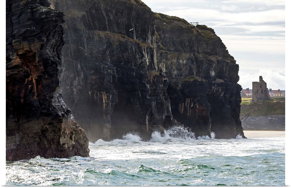 Dark straight cliffs with waves crashing into the rock with ruined castle turret in background, Ballybunion, County Kerry,...