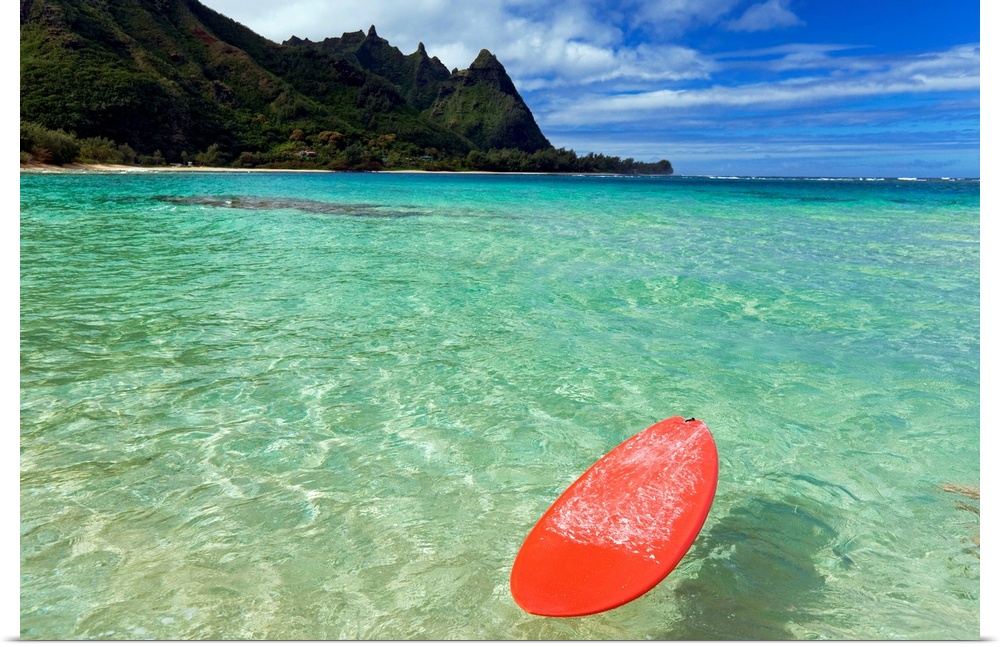 Photograph of bobbing surfboard in ocean with crystal clear waters and a small beach with tree covered mountains in the di...
