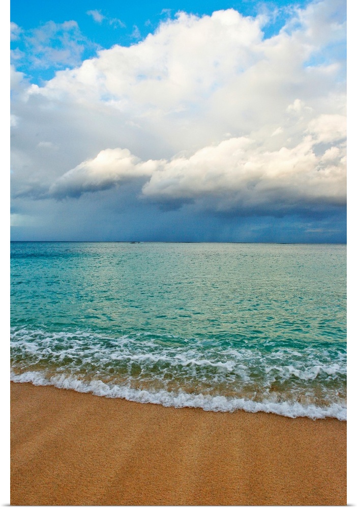 This photograph is taken on a beach in Hawaii of immense clouds that hang in the sky over a teal colored ocean.