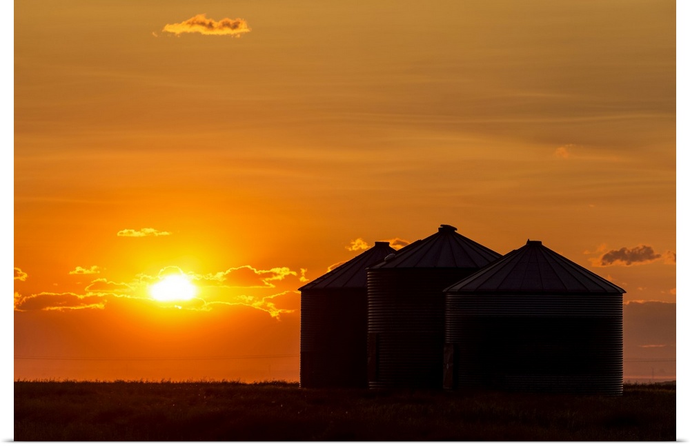 Silhouette of large metal grain bins at sunrise with orange sun rising over clouds; Alberta, Canada