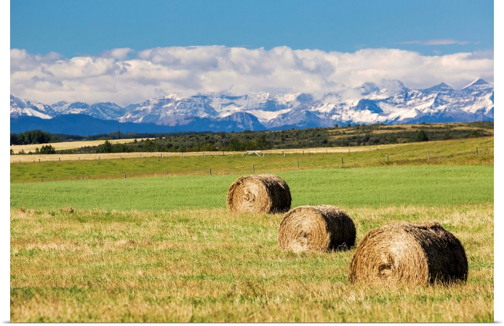 Three hay bales in a field with mountains in the background slightly snow covered and cloudy with blue sky, Alberta, Canada.