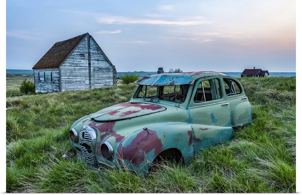 Vintage car sitting in the overgrown grass in a field with old buildings on a farmstead; Saskatchewan, Canada