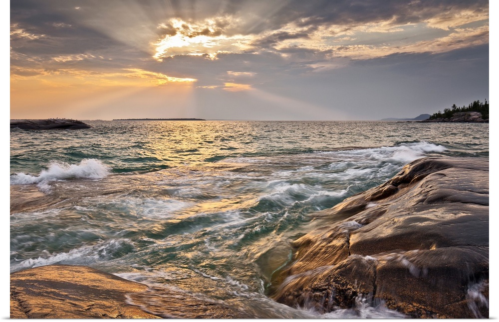 Burst of sun rays peeking out behind a cloud as waves lap on to a rocky shore.