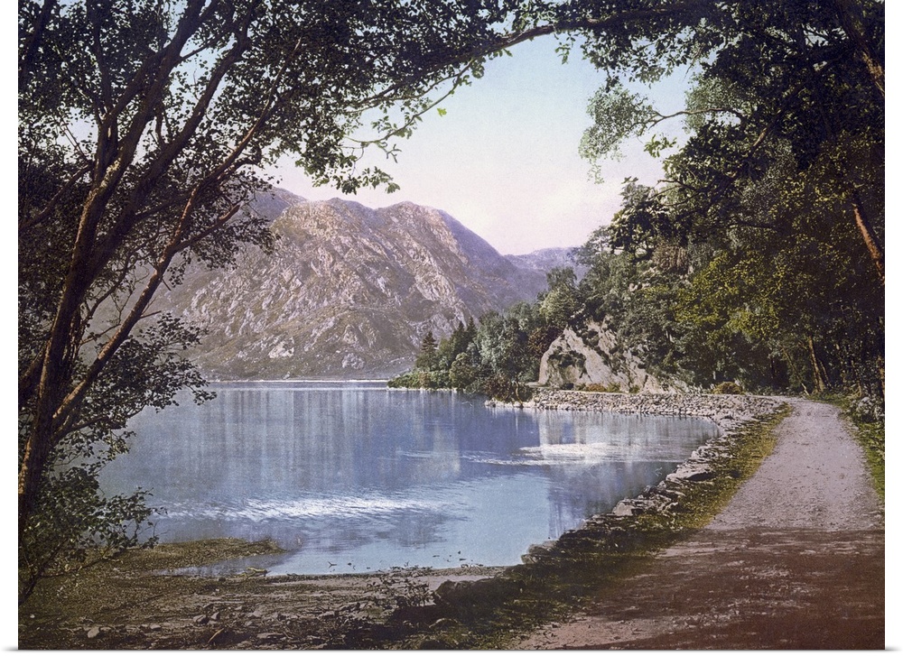 A vintage shot of a gravel path winding its way at the water's edge as leafy trees and cliffs loom overhead.