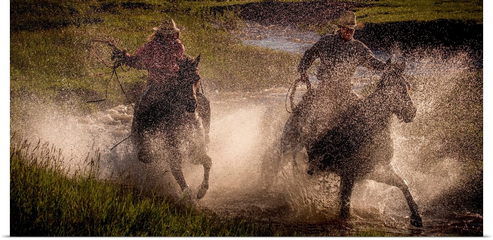 Photograph of two cowgirls splashing through a steam on horseback with their lassos out.