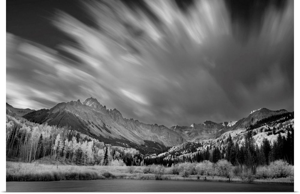 Black and white landscape photograph of a lake in front of a mountain range with blurred moving clouds in the sky.
