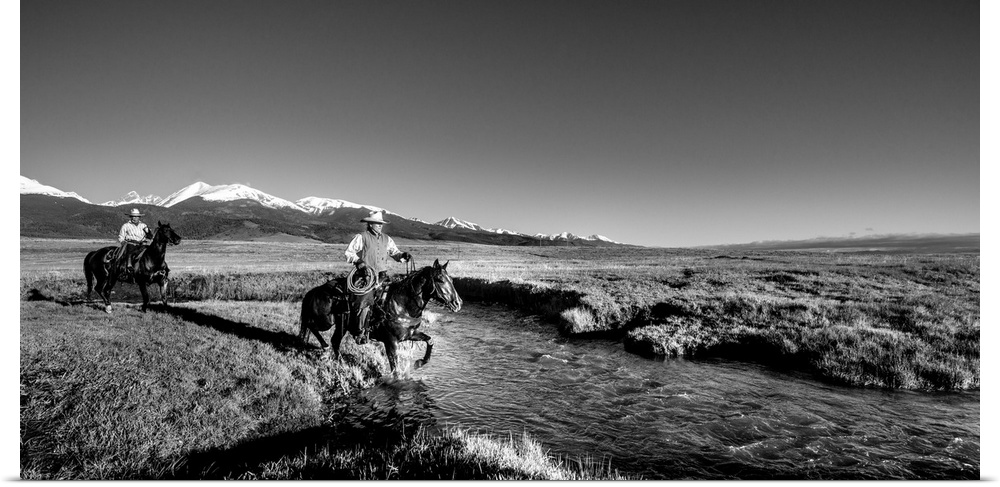 Black and white photograph of two cowgirls crossing a river on horseback with snow capped mountains in the distance.