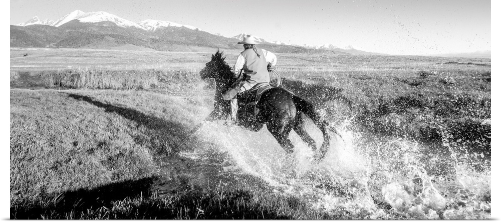 Action photograph of a cowgirl splashing across a river on horseback with snow capped mountains in the distance.