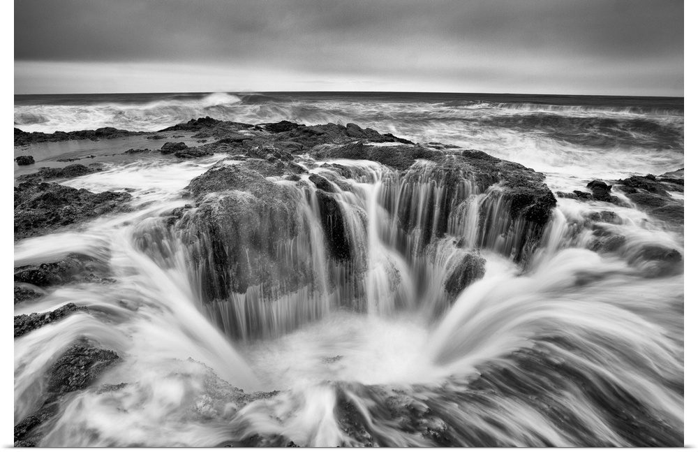 A black and white photograph of a well in the ocean surrounded by rushing water.