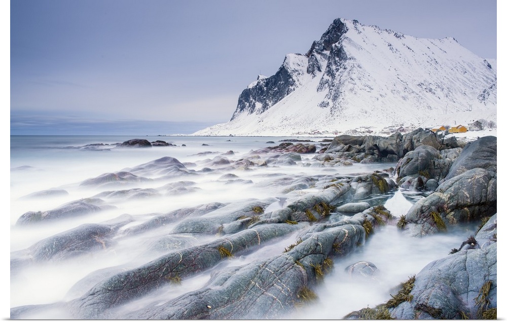 A photograph of a snow covered mountain seen from a rocky shoreline with motion blurred water caught in a long exposure.