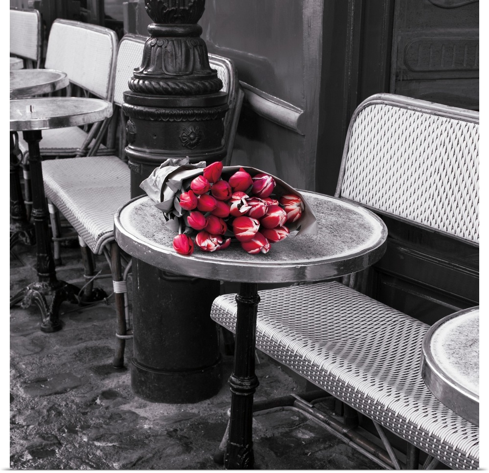 Square black and white photograph of sidewalk seating at a cafe with colored flowers on a table.
