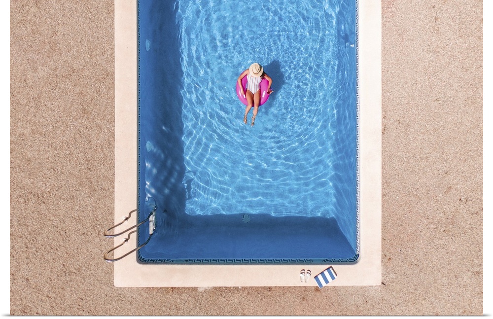 Zenith aerial view of a swimming pool in summer. Young girl in a swimsuit and hat floating with pink donut.