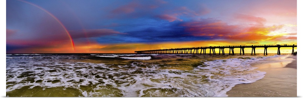 A dark blue sky with a purple sunset featuring a full rainbow. A long pier can be seen reaching into the ocean.