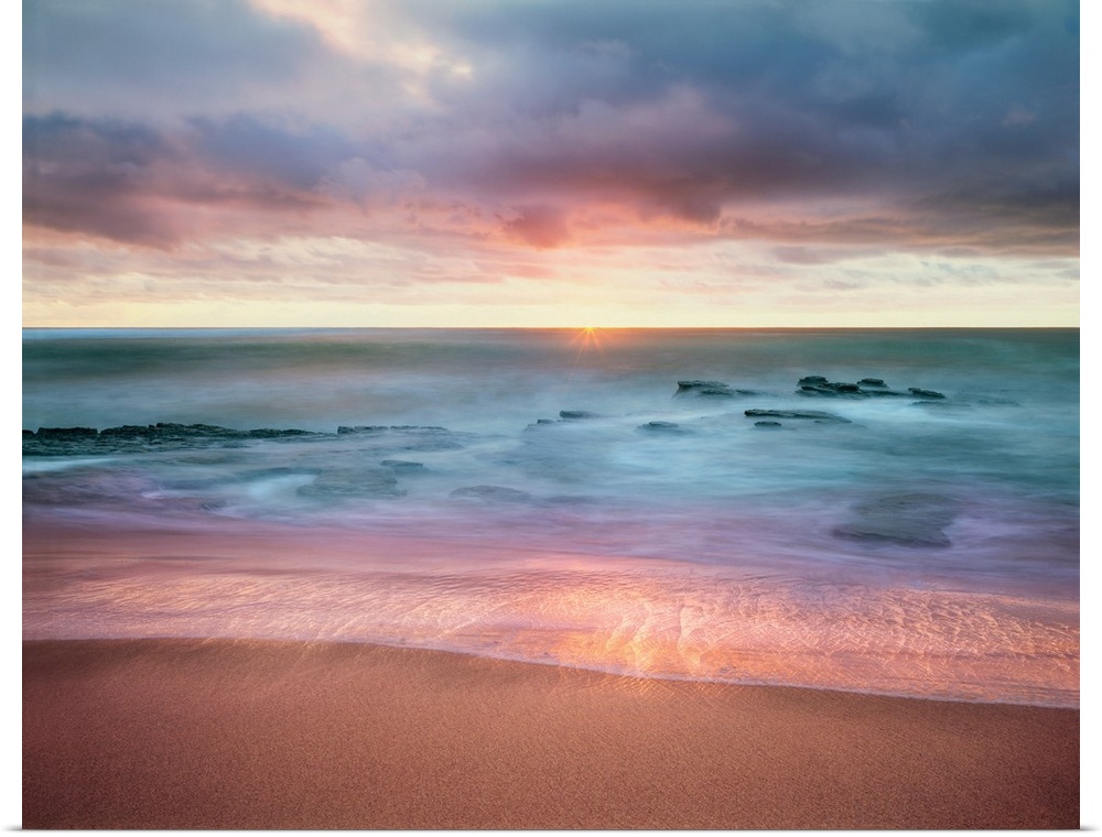 Colorful long exposure photograph a rocky ocean shore with the sun on the horizon in shades of blue, purple, and pink.