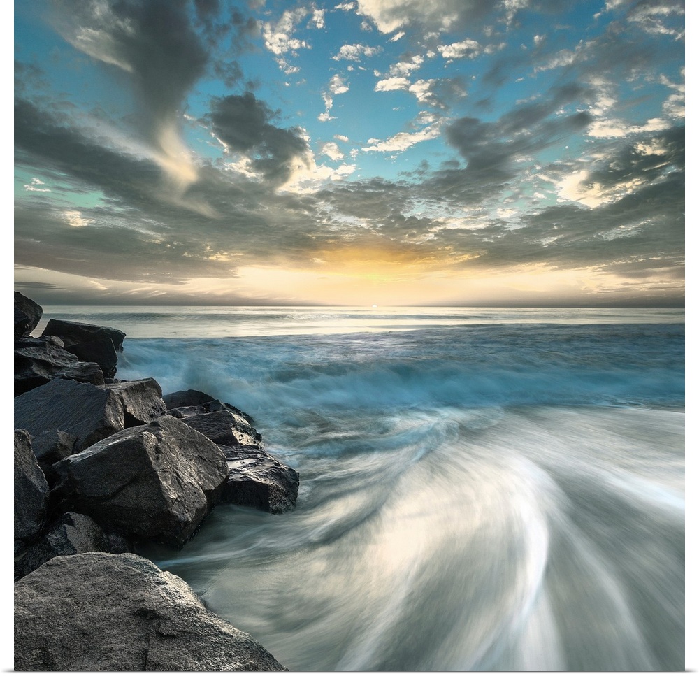 Long exposure photograph of ocean waves crashing on a rocky beach shore with a dramatic sky.