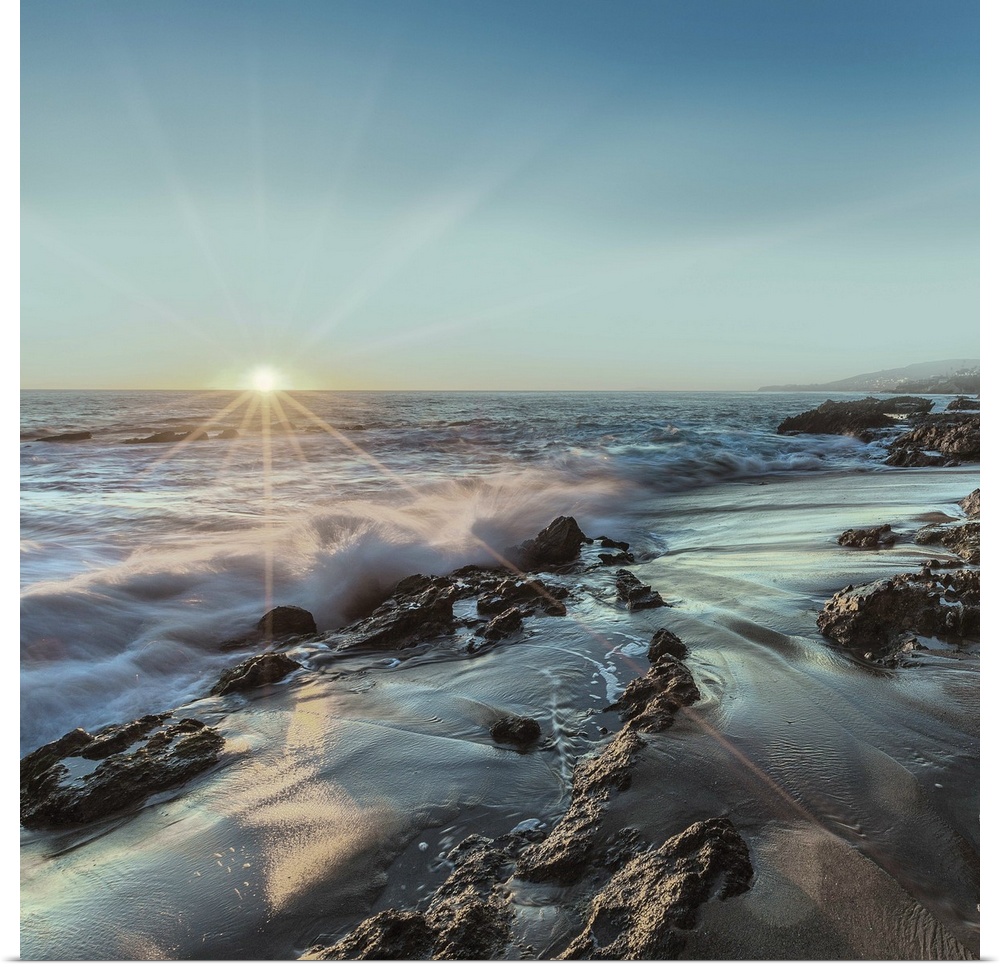 Square photograph of ocean waves crashing on a rocky shore with the sun setting on the horizon line at Laguna Beach.