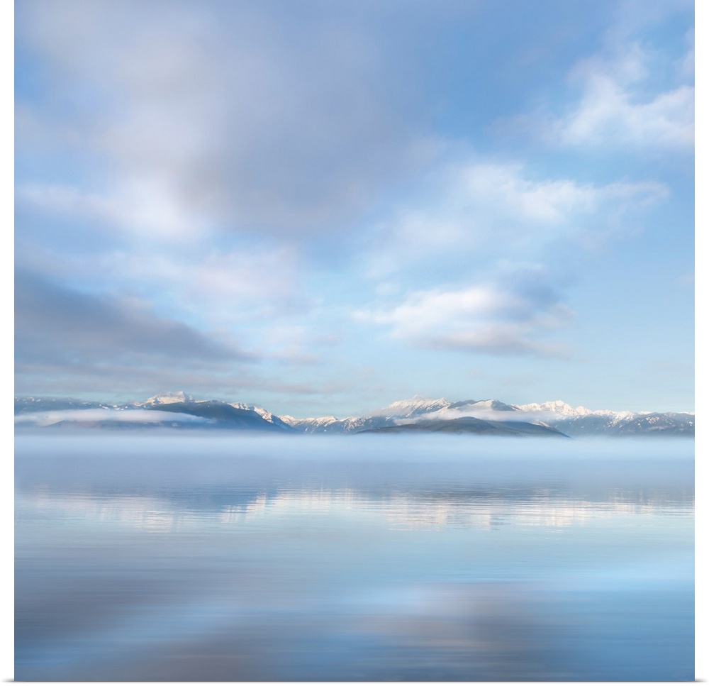 Square photograph of a snow capped mountain range in the distance with fluffy clouds above reflecting onto the ocean.
