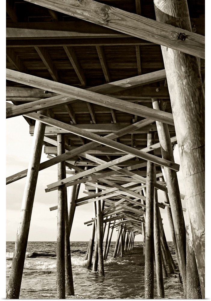 Big monochromatic photograph taken from beneath a large wooden pier examines the foundation of it while the ocean crashes ...