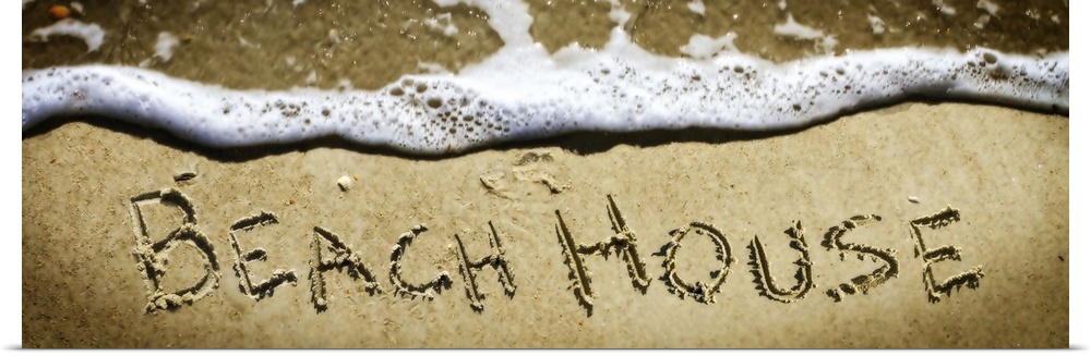The word "Beach House" drawn in the sand near the ocean water.