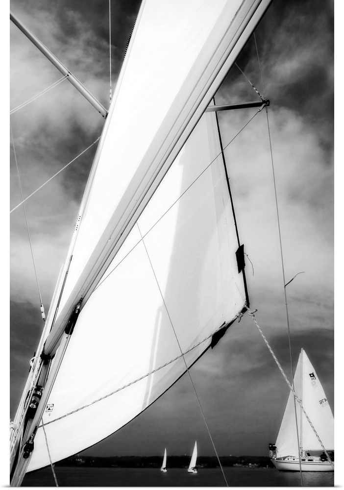 Giant, vertical, close up photograph of a sail blowing in the wind, in front of a cloudy sky.  Several smaller sailboats c...