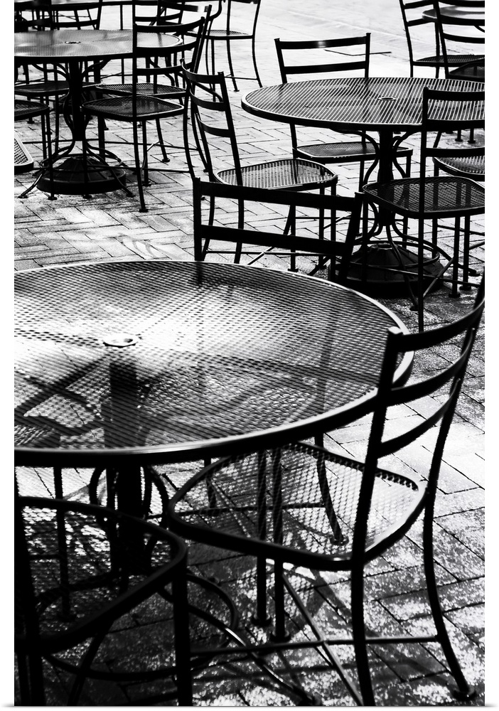 Black and white photograph taken of outdoor tables and chairs that sit on a brick sidewalk.