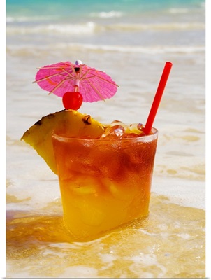 A mai tai garnished with pinapple and a cherry, sitting in shallow water on the beach.