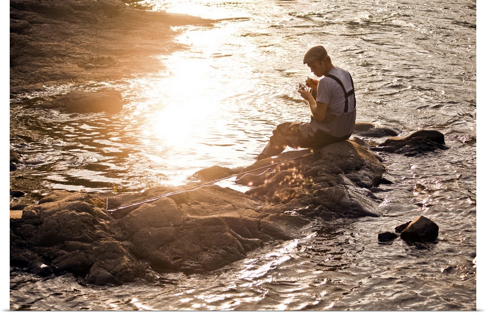 A young fisherman working on his rod and reel next to a river at sunset.