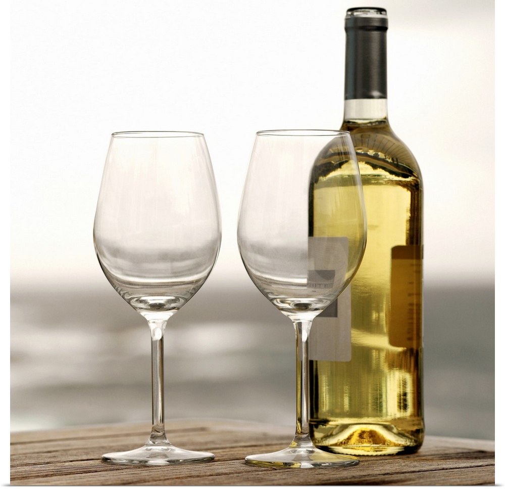 Square photograph on a large canvas of two empty wine glasses sitting on a wooden table, along with a full bottle of white...