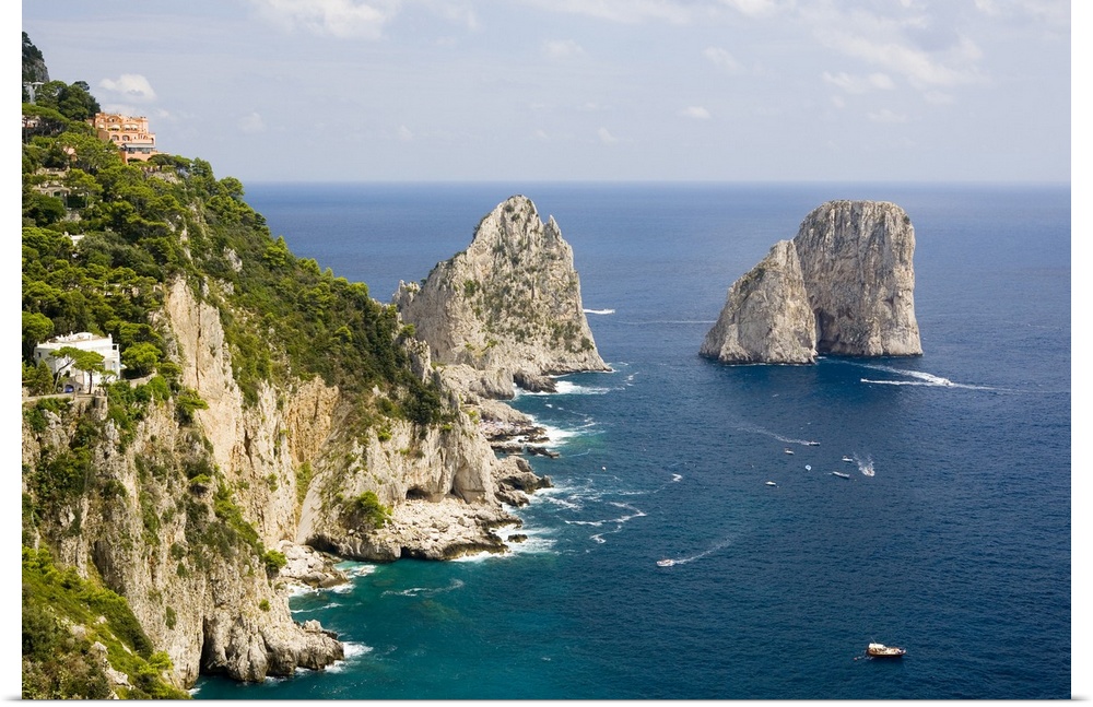 Large rock formations stand in the water next to an immense cliff off the Italian coast.
