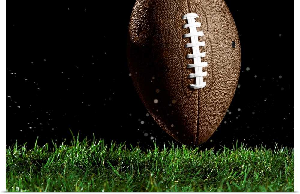 Giant landscape photograph of a football in a vertical position as it moves above the wet grass with specks of water flyin...