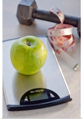 Green apple on weight scale, tape measure and exercise weight in background