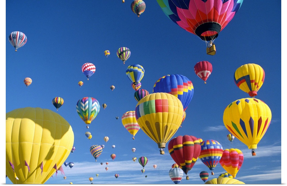 Big, horizontal photograph of many brightly colored hot air balloons with varying patterns, floating against a blue sky.