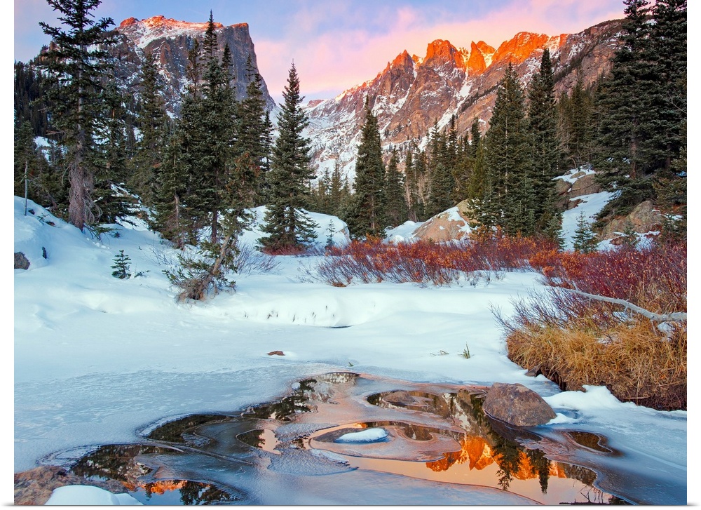 Photograph of a little creek by snow-covered ground and trees with the Rocky Mountains in the background in Rocky Mountain...