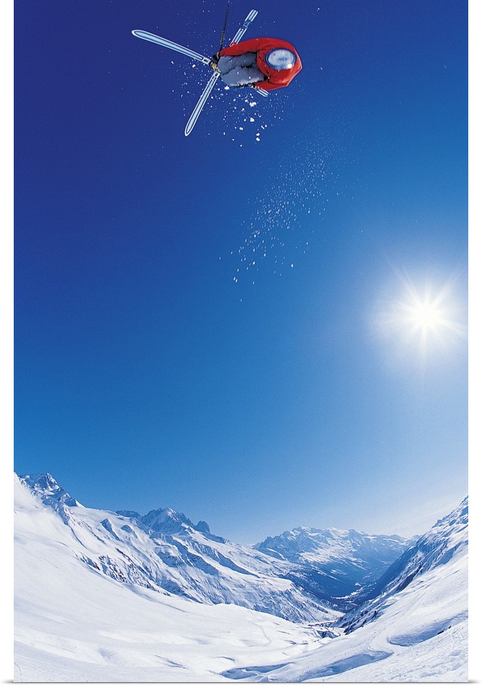 Vertical photo of a man crossing his skis on as he flies through the air as the sun shines down on the snow covered mounta...