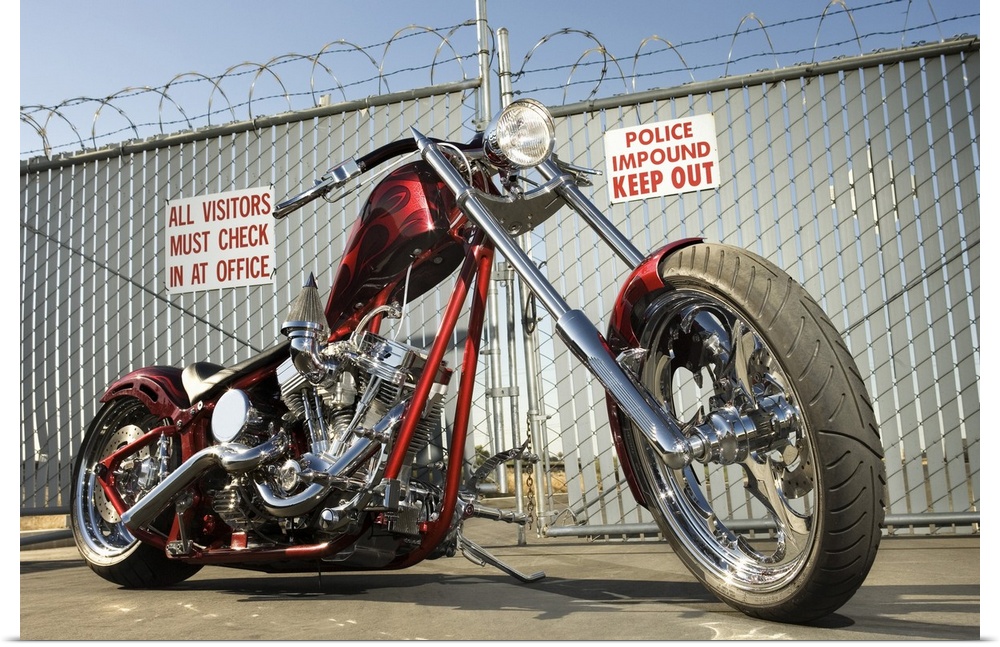 A chrome and red motorcycle sits on the the conrete pavement infront of a barbed wire fence surrounding an impound lot.
