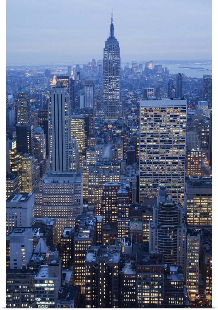 Large vertical photograph of NYC taken at dusk with the Empire State Buiding center and many other buildings lit up surrou...