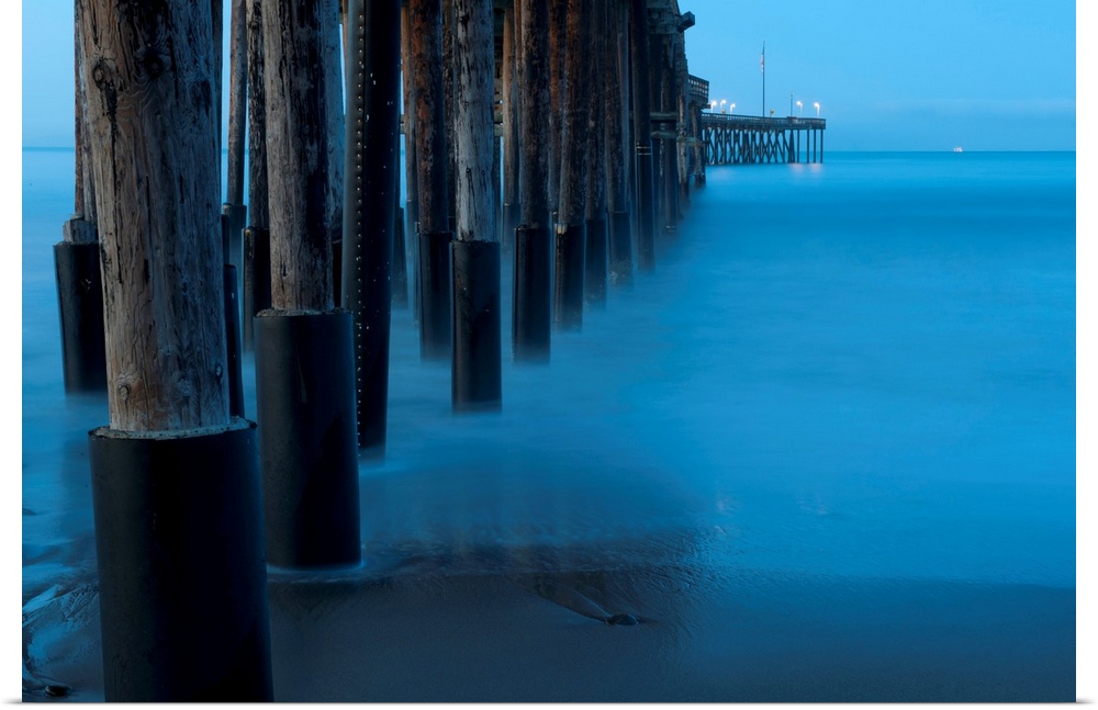 Large print of tall wooden pier pillars holding up a pier leading into the ocean from the shore.