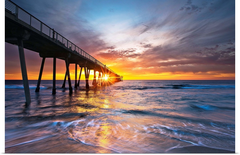 Sun intersects pier at sunset with colorful waves and sky.