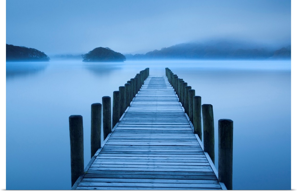 A photograph is taken looking down a pier that stands over a body of water and looks out onto rock formations which are ba...