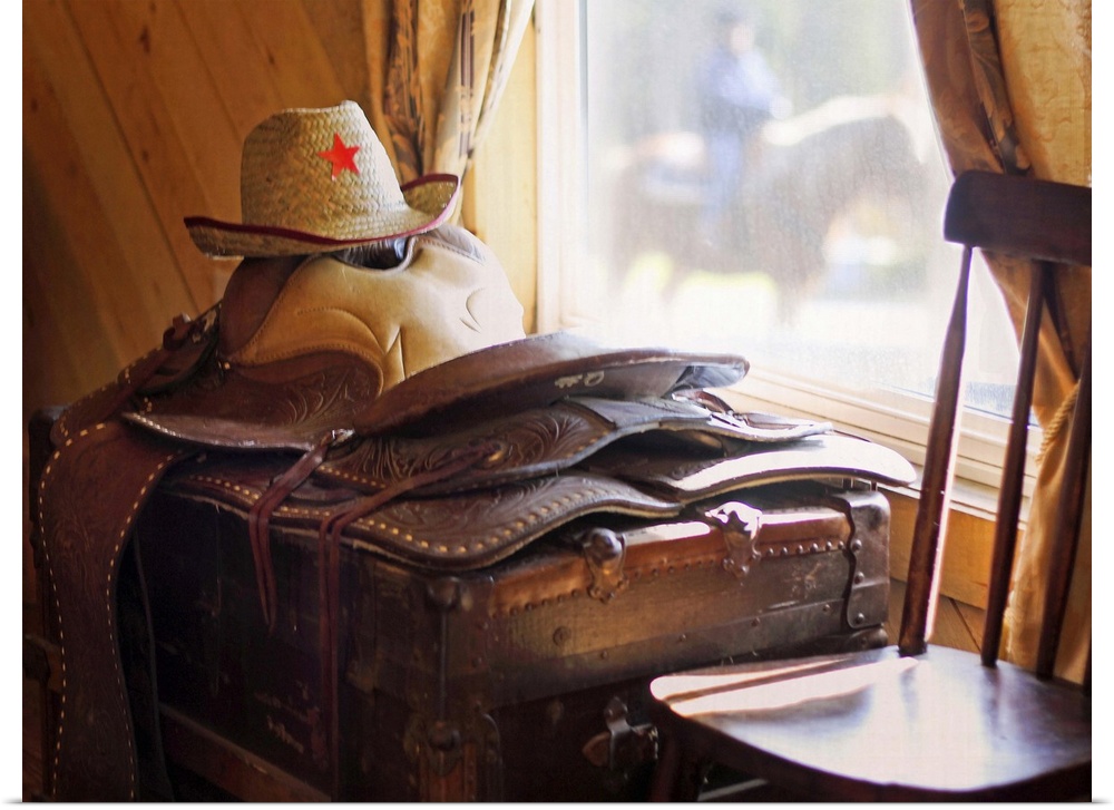 Leather saddle and cowboy hat resting on antique trunk catching the light through a window with out-of-focus horse and rid...