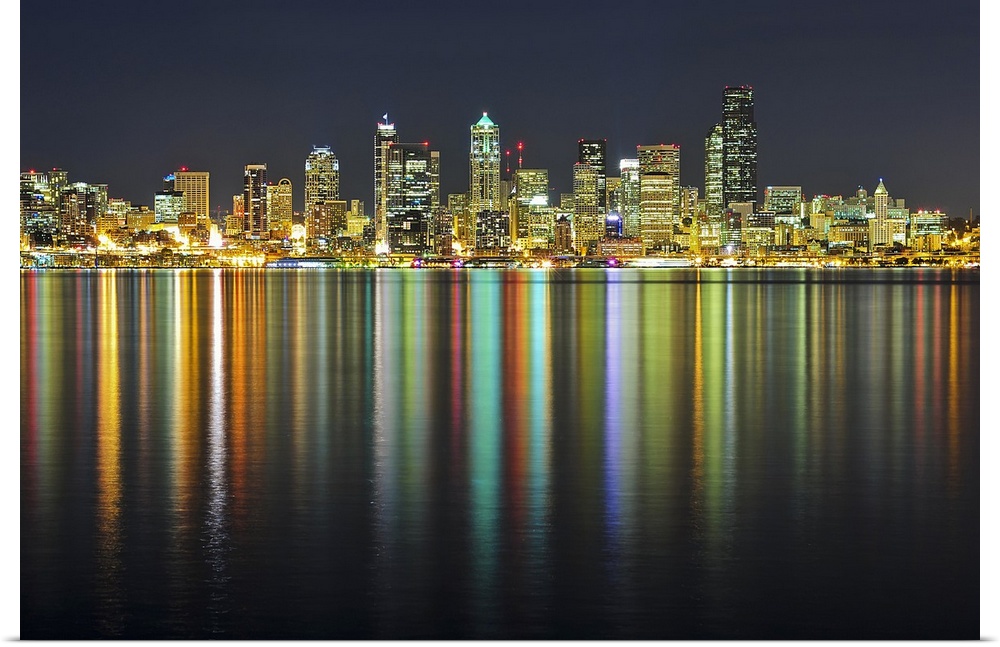 Large photograph taken of the Seattle skyline at night with the buildings lit up and reflecting in the water below.