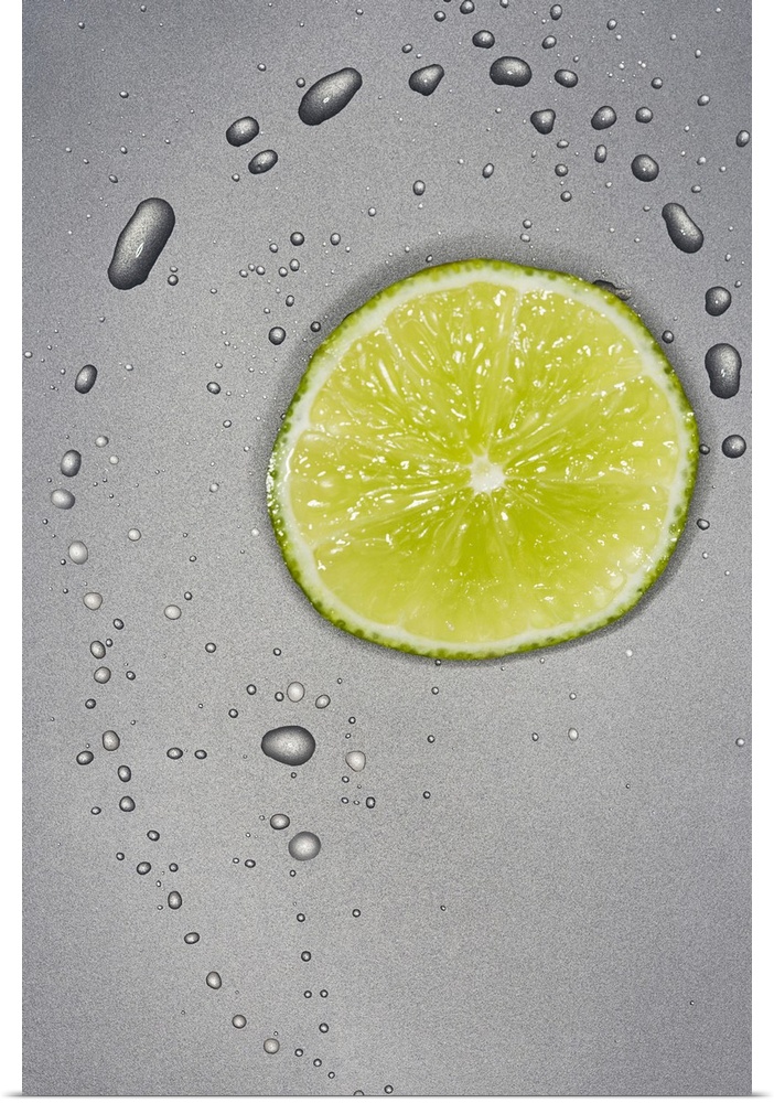 This photograph is taken of a slice of a lime with water droplets surrounding it on a grey surface.