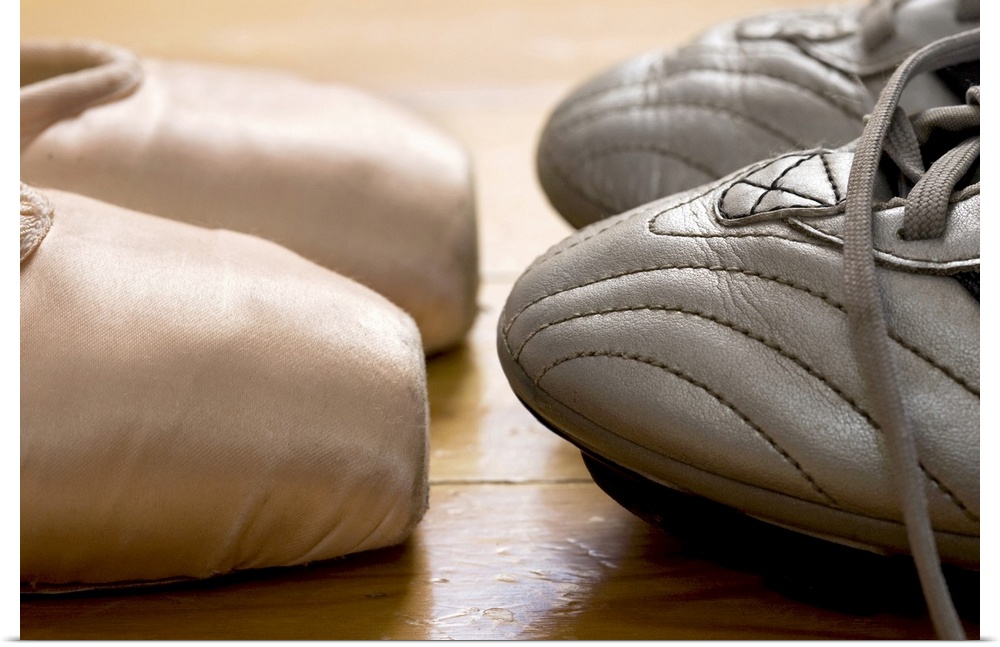 Photograph of the tips of ballet point shoes facing the tips of football cleats on a hardwood floor.