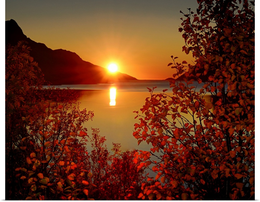 Big photograph emphasizes the sun as it begins to set over a mountain and then reflect over a calm body of water in Norway...