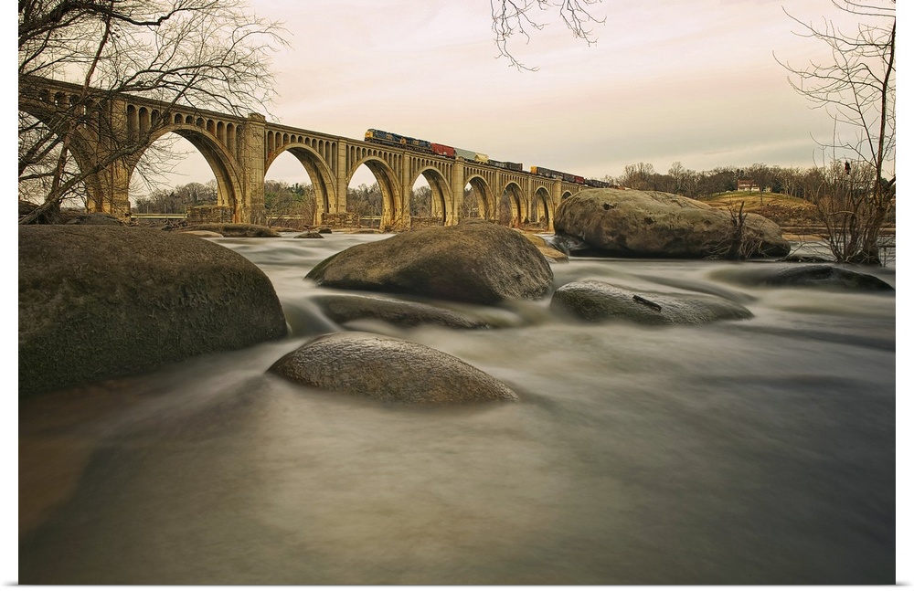 Large rocks sit on the banks for a river with a multi arched bridge spanning the river and a train running on top of the b...