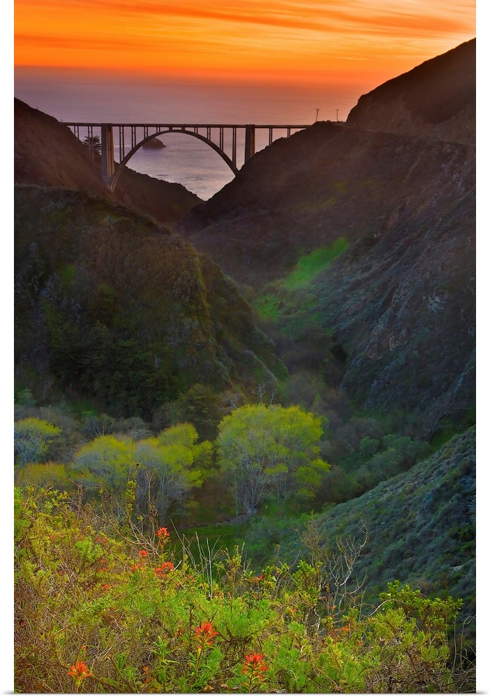 Portrait oriented photo of the Big Sur bridge at sunset with rolling hills in the foreground and ocean in the background.