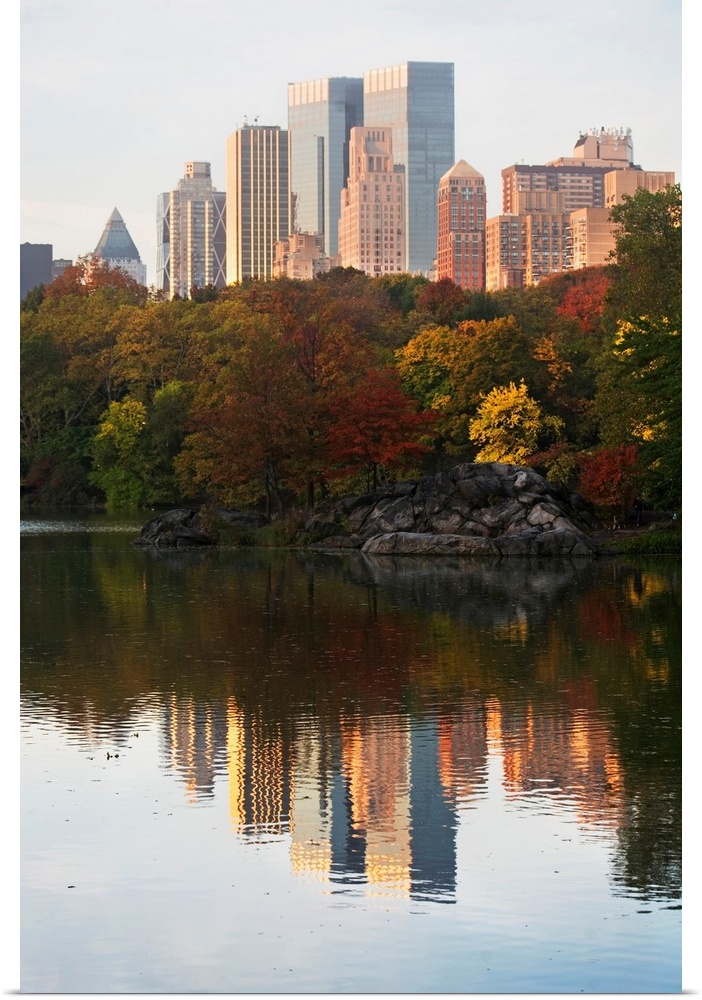 This is a vertical photograph of autumn trees and skyscrapers reflecting in the calm waters of this city park pond.
