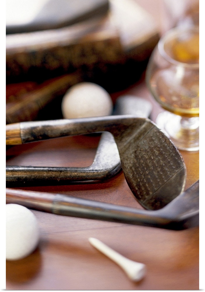 Antique golf club heads are photographed laying on a table with golf balls, a tee and a glass of liquor sitting beside them.