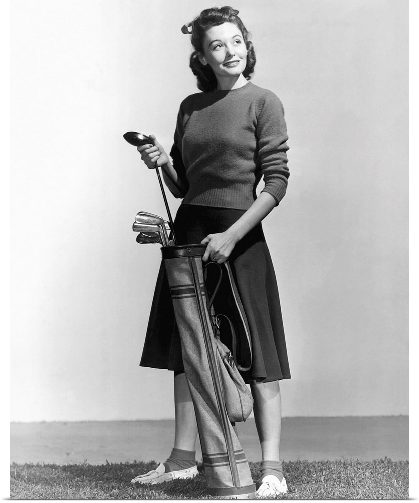 Woman holding a golf club and a golf bag, and a golf ball is lying on the ground. Full length photograph. Ca. 1940s-1950s.